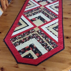 May include: A red, black and gold table runner with a geometric pattern. The runner has a black border and a red border with a black trim.