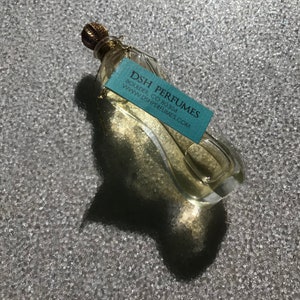 Op de afbeelding: Een doorzichtige glazen fles met een gouden dop en een blauw etiket met de tekst "DSH PERFUMES, BOULDER, CO 80304, WWW.DSHPERFUMES.COM".