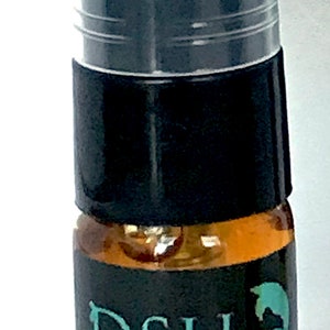 May include: A small perfume bottle with a black cap and a clear plastic spray top. The bottle has a dark label with the text "DSH Perfumes" and a teal leaf design. The liquid inside is amber-colored.