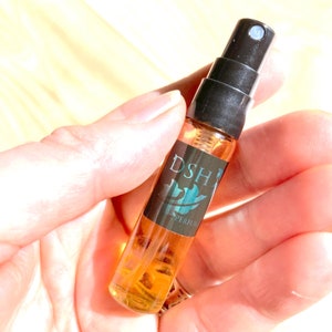 Op de afbeelding: Een klein, doorzichtig glazen flesje met een zwarte spuitdop en een zwart etiket waarop "DSH" staat met een turquoise bladerenontwerp. Het etiket vermeldt ook "PERFUME".
