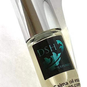 Op de afbeelding: Een doorzichtige glazen fles met een zilveren dop en een zwart etiket met de tekst "DSH Perfumes" en "Aqua Calma oil ess DSHPerfumes.com".