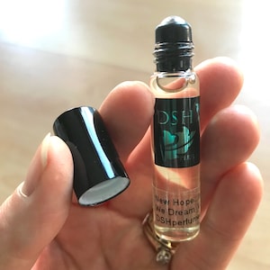 May include: A black and clear glass roller bottle with a black cap. The bottle has a label with the text "DSH Perfumes" and the words "New Hope... We Dream..." in white text.