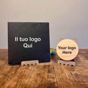 Può includere: Due cartelli in legno con testo nero su un tavolo in legno. Il cartello a sinistra è una lavagna nera con il testo "Il tuo logo Qui". Il cartello a destra è un cartello rotondo in legno con il testo "Your logo Here".
