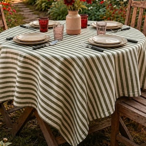 Op de afbeelding: Een ronde houten tafel gedekt voor een maaltijd in de buitenlucht, bedekt met een groen-wit gestreept tafelkleed. De tafel is gedekt met borden, bestek en glazen. Een vaas met bloemen staat in het midden. Twee houten stoelen staan aan weerszijden van de tafel.