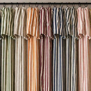 May include: A collection of striped curtains in various colors, including green, gray, orange, burgundy, navy, and pink. The curtains are hanging from a black rod, showcasing a range of vertical stripe patterns. The fabric appears to be linen or a similar natural material.