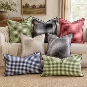 Pode incluir: Uma coleção de almofadas decorativas com padrão xadrez em várias cores, incluindo verde, preto, vermelho, azul e cinza. Cada almofada apresenta uma borda contrastante. As almofadas estão dispostas em um sofá branco.