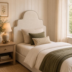 Op de afbeelding: Een gestoffeerd hoofdeinde met een geschulpte bovenrand in een lichtbeige stof. Het bed is voorzien van een groot beige kussen, een donker olijfgroen rechthoekig kussen, een kleiner kussen met patroon en een doorgestikte olijfgroene bedloper.