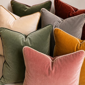 May include: A collection of square velvet throw pillows in various colors including olive green, cream, charcoal gray, rust red, mustard yellow, and dusty rose. Each pillow features a contrasting piped edge trim along the seams of the fabric.