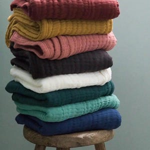 May include: A pile of folded, textured blankets in shades of burgundy, mustard, dusky pink, black, white, teal, sea green, and navy. The blankets are stacked on a small, round wooden stool.