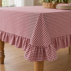 May include: A rectangular tablecloth featuring a red and white gingham check pattern. The fabric drapes over the edges of a wooden table and includes a gathered ruffle trim along the entire bottom border, creating a layered and textured appearance.