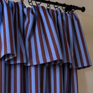 May include: A set of curtains featuring vertical stripes in alternating shades of light blue and dark brown. The fabric is gathered at the top and suspended from a black metal curtain rod with circular rings, creating a series of soft folds.