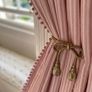 May include: Pink and white striped curtains with a gold-colored rope-style tieback. The curtains have a pom-pom trim along the edge. The tieback features a bow design with tassel ends, adding a decorative touch to the window treatment.