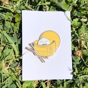 May include: A gold-colored enamel pin featuring a cartoon dumpling in a yellow bamboo steamer with a lid. Two gold chopsticks are placed next to the steamer. The pin is set on a white card against a background of green grass.