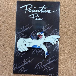 Könnte beinhalten: Ein weiß-blauer Emaille-Pin einer Fabelwesen-Figur mit ausgebreiteten Flügeln, offenem Mund und blauen Akzenten. Der Pin befindet sich auf einer schwarzen Rückkarte mit dem Schriftzug "Primitive Pins" in weißer Schrift.