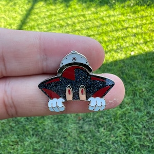 May include: A distinctive enamel pin depicting a black creature with red details, white gloves, and a white hat. The pin has a glittery black body and gold-coloured metal accents. The design is playful and visually appealing.