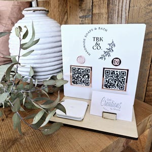 May include: A white display stand featuring the text "TRK & CO. HANDMADE SOAPS & BATH" and two QR codes. A business card with the text "Little Creations by Shannie" is displayed. The stand is on a wooden surface.
