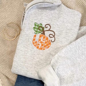 May include: A light gray quarter-zip sweatshirt featuring an embroidered orange and white gingham pumpkin design with a green gingham leaf. The pumpkin graphic includes the letters LSN in a bold, block font. Two gold hoop earrings are visible.