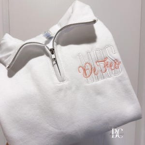 May include: White quarter-zip sweatshirt with a silver zipper. The sweatshirt features the embroidered text "MRS De Feo" in a combination of white and light pink thread. The garment is folded, showcasing the embroidered details.