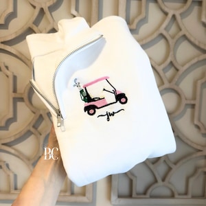 May include: A white quarter-zip sweatshirt featuring an embroidered design of a pink golf cart with a green golf bag containing clubs. Below the golf cart, the letters jw are embroidered in a black cursive script font on the front chest area.