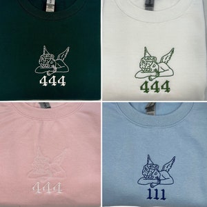 May include: Four sweatshirts in different colors: dark green, white, pink, and light blue. Each sweatshirt features an embroidered design of an angel with the number "444" or "111" below. The angel and numbers are in contrasting colors.