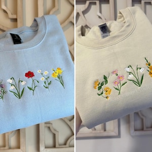 May include: Two crewneck sweatshirts, one light blue and one cream, each embroidered with a row of colorful flowers. The light blue sweatshirt features purple, white, red, and yellow flowers. The cream sweatshirt has yellow, pink, and white flowers.