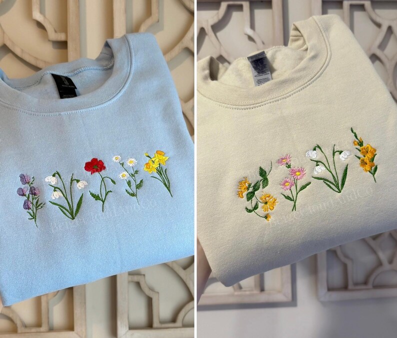 May include: Two crewneck sweatshirts, one light blue and one cream, each embroidered with a row of colorful flowers. The light blue sweatshirt features purple, white, red, and yellow flowers. The cream sweatshirt has yellow, pink, and white flowers.