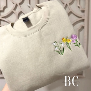 May include: A beige crewneck sweatshirt with embroidered flowers. The floral design includes white, yellow, and purple blossoms with green stems and leaves. The sweatshirt has a ribbed neckline and a tag on the back.