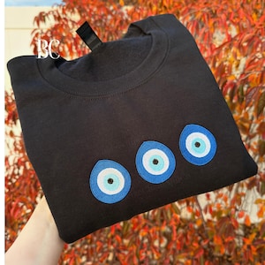 May include: Black crew neck sweatshirt with three embroidered blue and white evil eye designs. The sweatshirt is folded, showcasing the eye symbols. The background features fall foliage.