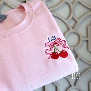 May include: A light pink sweatshirt with a crew neck. The sweatshirt features an embroidered design of two red cherries with a pink bow and the initials "LS" in blue. The design is on the upper left sleeve.