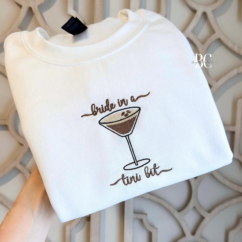 White crewneck sweatshirt folded neatly, featuring a small embroidered martini glass with the phrase “bride in a tini bit” in soft neutral thread on the center chest, creating a minimal and elegant bridal design.