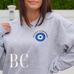 May include: Gray sweatshirt with a blue evil eye design and the text "Protect Your Energy". The sweatshirt also has the text "BC Berge Cloud And Co." on the bottom.