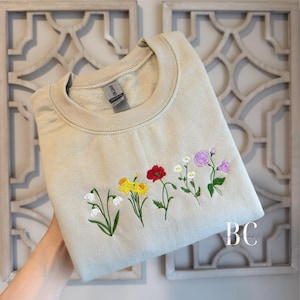May include: Beige crewneck sweatshirt with embroidered flowers in white, yellow, red, and purple. The floral design is stitched across the chest. The sweatshirt is held up by a hand.