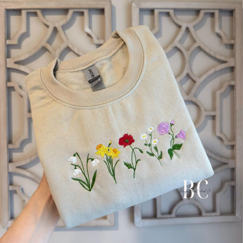 May include: Beige crewneck sweatshirt with embroidered flowers in white, yellow, red, and purple. The floral design is stitched across the chest. The sweatshirt is held up by a hand.