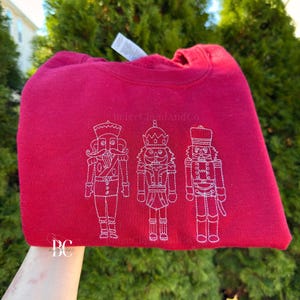May include: A red sweatshirt with a white embroidered design of three nutcrackers. The text "BeigeCloudAndCo" is above the nutcrackers. The sweatshirt is folded and held up.