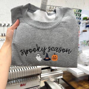 May include: A heather gray crewneck sweatshirt featuring black embroidered text that reads spooky season. Below the text are three small embroidered icons: a white ghost, a black witch hat with an orange band, and an orange jack-o'-lantern.
