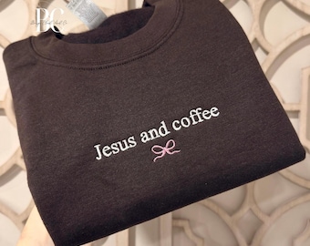 May include: A dark brown sweatshirt with the embroidered text "Jesus and coffee" in white, accompanied by a small pink bow. The sweatshirt is a crew neck style.