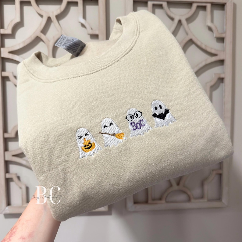 May include: A beige crewneck sweatshirt featuring an embroidered design of four white ghosts. The first ghost holds an orange pumpkin, the second holds a brown broom, the third wears black glasses with the purple text "Boo" below, and the fourth wears a black bat mask.