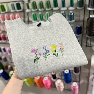May include: A heather gray sweatshirt featuring an embroidered floral design. The embroidery includes purple, blue, yellow, and pink flowers. The sweatshirt is folded, and the image is taken indoors.