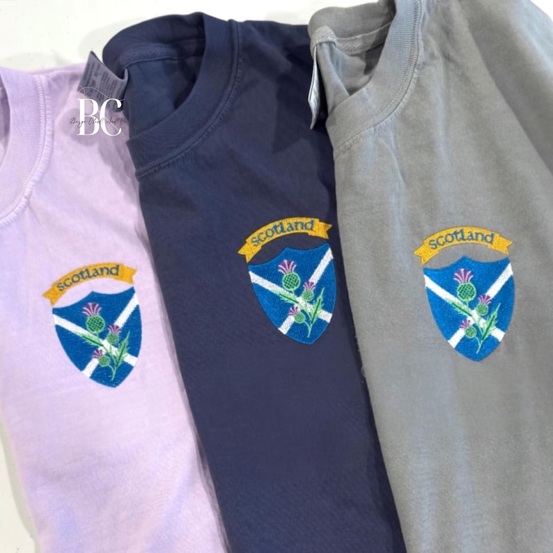 Three Comfort Colors t-shirts in shades orchid, navy and grey are shown next to eachother. Each tshirt features a colorful embroidered Scotland crest on the upper left chest. The crest includes the Scottish flag, purple thistles, and a gold banner reading “Scotland.”