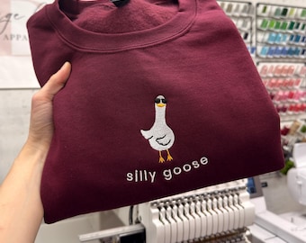 May include: A maroon crewneck sweatshirt featuring an embroidered graphic of a white goose wearing black sunglasses. Below the goose, the text "silly goose" is embroidered in white thread. The sweatshirt has a ribbed collar and long sleeves.