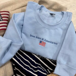 May include: A light blue crewneck sweatshirt featuring dark blue embroidered text that reads 'loves Jesus & America too' positioned above a small, detailed embroidered American flag with red, white, and blue stripes and stars.