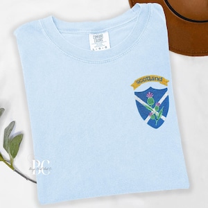 A folded light blue Comfort Colors t-shirt featuring a colorful embroidered Scotland crest on the upper left chest. The crest includes the Scottish flag, purple thistles, and a gold banner reading “Scotland.”