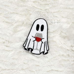 May include: Embroidered patch featuring a white ghost holding a wine glass filled with red liquid. The ghost has black oval eyes and a black outline. The wine glass has a silver stem and a red wine. The patch is on a white, textured background.
