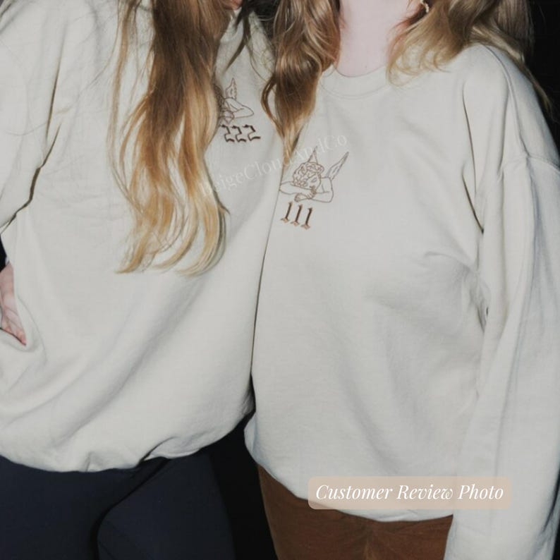 May include: Two beige sweatshirts with long sleeves. Each sweatshirt has a graphic of an angel with the numbers 222 and 111 embroidered in brown thread. The text "Angel Cloud Co" is also visible.