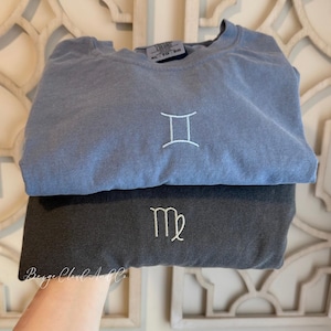 May include: A blue crewneck sweatshirt featuring an embroidered white Gemini zodiac symbol and a dark gray crewneck sweatshirt featuring an embroidered white Virgo zodiac symbol. Both garments have a visible size tag reading XL TG EG.