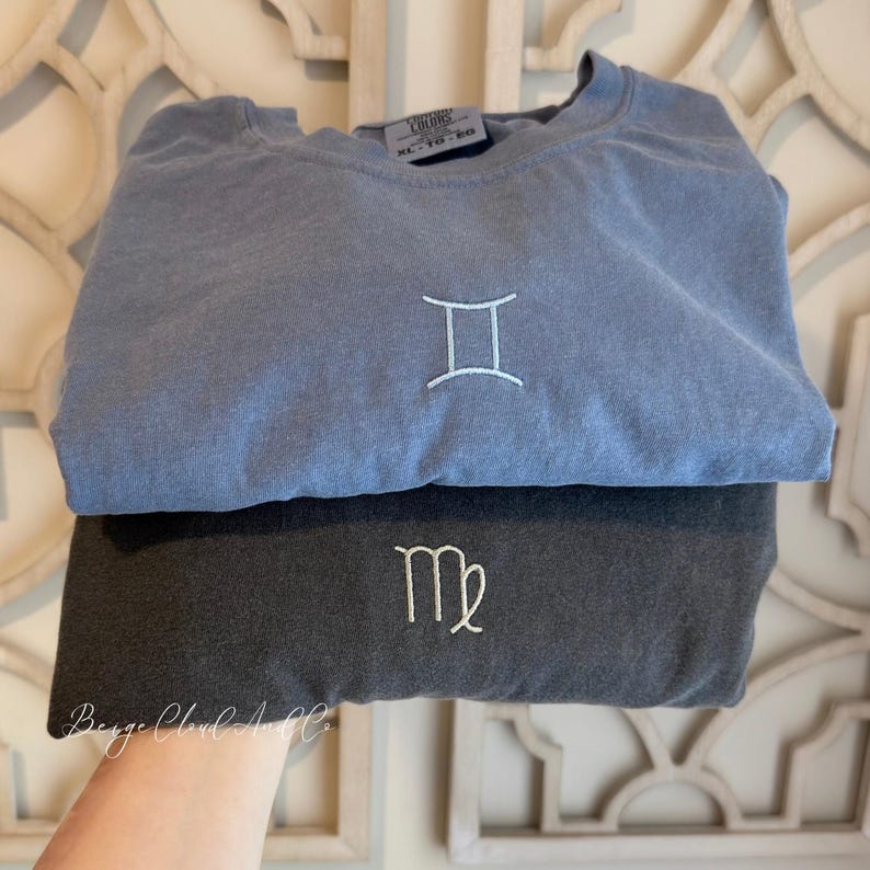 May include: A blue crewneck sweatshirt featuring an embroidered white Gemini zodiac symbol and a dark gray crewneck sweatshirt featuring an embroidered white Virgo zodiac symbol. Both garments have a visible size tag reading XL TG EG.