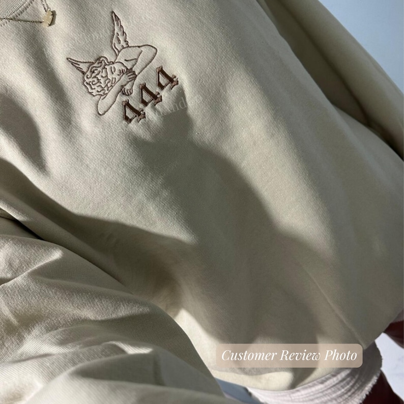 May include: Cream-colored sweatshirt featuring an embroidered design of an angel and the number "444" in brown thread. The sweatshirt has a relaxed fit and a simple, minimalist aesthetic. The image is a customer review photo.