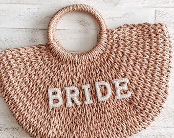 May include: A straw tote bag with a round handle and the word "BRIDE" spelled out in white pearl letters.