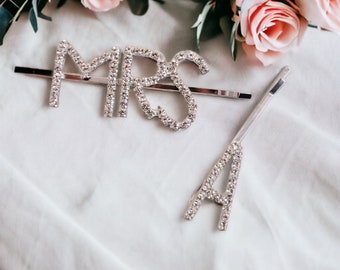 Könnte beinhalten: Zwei silberne Haarklammern mit Strasssteinen. Eine Klammer buchstabiert "MRS" und die andere Klammer buchstabiert "A".