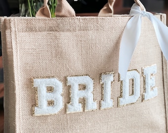 May include: A beige burlap tote bag with white fabric letters spelling "BRIDE" in a varsity style font. The letters have gold glitter accents. A white satin ribbon is tied to the handles.